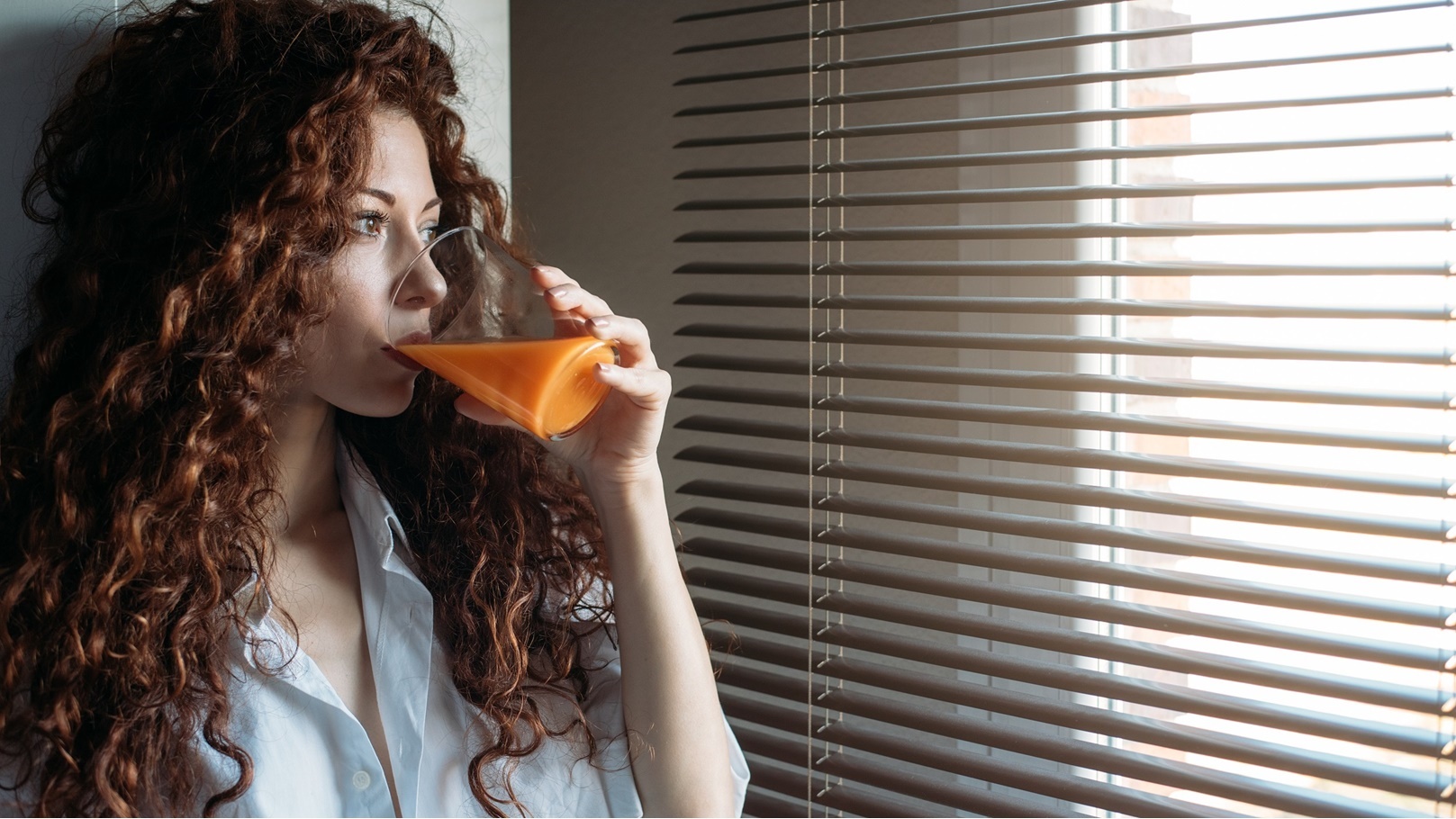 woman-standing-by-a-windows-blind-drinking-orange-2022-03-04-06-11-22-utc